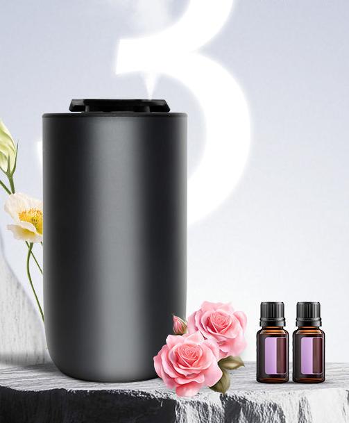 WSNUEEE A613 waterless aroma diffuser in matte black standing on a stone plinth beside pink roses and two 15 mL essential oil bottles, styled for home and car use